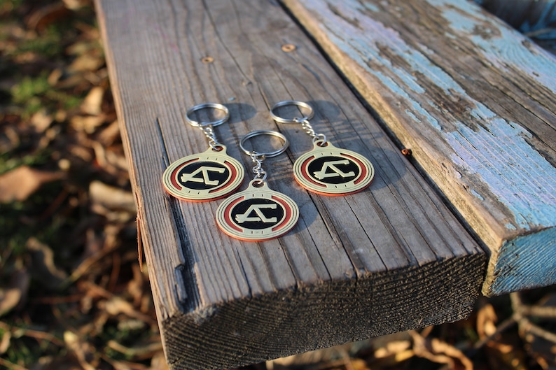 May include: Three keychains with a gold-colored circular pendant featuring a black letter "A" inside a red and gold border. The keychains are on a wooden surface.