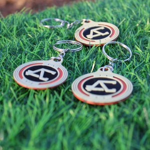 May include: Three keychains with a silver ring and a circular pendant. The pendant has a gold border, a red inner ring, and a black center with a white "A" and "C" design. The keychains are on green grass.