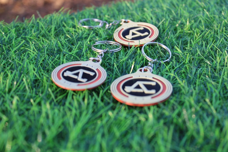 May include: Three keychains with a silver ring and a circular pendant. The pendant has a gold border, a red inner ring, and a black center with a white "A" and "C" design. The keychains are on green grass.