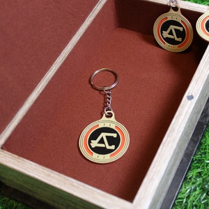 May include: A collection of gold-colored keychains and pendants with a black and orange design, displayed inside a wooden box with a brown interior. The keychains have a circular shape with a stylized letter "A".