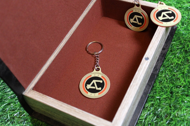 May include: A collection of gold-colored keychains and pendants with a black and orange design, displayed inside a wooden box with a brown interior. The keychains have a circular shape with a stylized letter "A".