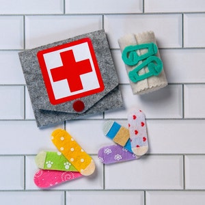 May include: A grey felt first aid kit with a red cross on the front. The kit is surrounded by colourful felt bandages with different patterns.