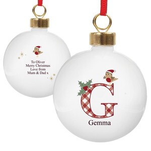 May include: Two white Christmas baubles with gold-coloured tops and red ribbons. One features a red and white checked letter "G" with a bird wearing a Santa hat. The other has the text "To Oliver Merry Christmas Love from Mum & Dad x".
