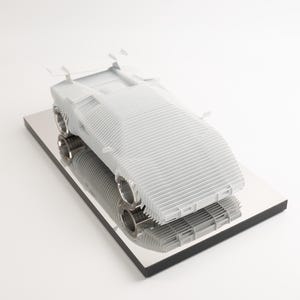 May include: A light grey model car with a ribbed design, featuring a spoiler and chrome exhaust pipes. The car sits on a reflective, black-edged base, creating a mirrored effect. The design suggests a modern, aerodynamic aesthetic.