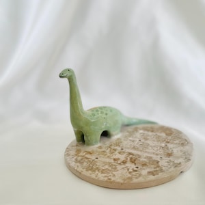 Può includere: Statuetta di dinosauro in ceramica verde, posizionata su una base rotonda beige strutturata. Il dinosauro ha un lungo collo e un viso sorridente. La base presenta diversi piccoli fori.