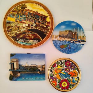May include: Four decorative souvenir magnets. One round magnet features a bridge and building with the text "Koprivshtitsa". Another round magnet depicts a harbor scene. A rectangular magnet shows a bridge and building with the text "Budapest - Lanchid". A round magnet has a bird and floral design.