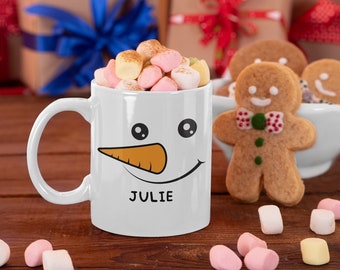 May include: A white ceramic mug featuring a printed snowman face with black eyes, an orange carrot nose, and a black curved smile. The name JULIE is printed in black capital letters below the smile. The mug is filled with colorful marshmallows.