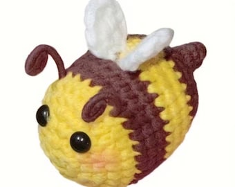 May include: A crocheted bee plush toy featuring a rounded body with alternating horizontal stripes of yellow and dark brown yarn. The bee has two small white crocheted wings on its back, two dark brown antennae, and two round black plastic eyes.