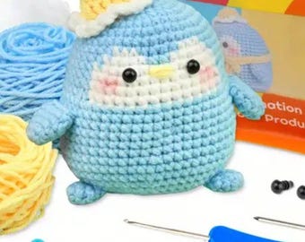 May include: A crochet kit featuring a light blue and white penguin plush with a yellow hat, accompanied by light blue and yellow yarn skeins, a blue crochet hook, and several colorful plastic stitch markers on a white surface.