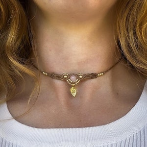 Puede incluir: Un collar gargantilla de macramé hecho a mano con una piedra preciosa rosa central y un colgante de hoja dorada. El collar está hecho con cordón marrón y cuentas doradas. La gargantilla se lleva alrededor del cuello.