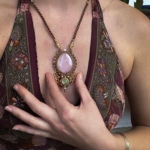 May include: A handcrafted necklace featuring a large, teardrop-shaped rose quartz pendant. The pendant is encased in a gold-colored, intricately woven setting, with a smaller green stone below. The necklace has a brown cord.