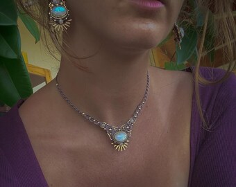 May include: A silver necklace with a central oval-shaped, light blue stone, surrounded by a gold sunburst design. Matching earrings are also visible. The jewelry is set against a purple long-sleeved top.