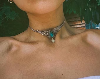 May include: A macrame choker necklace featuring a central oval-shaped teal gemstone set in a woven tan cord pattern. The design includes a small, dangling metallic charm suspended from the bottom center of the intricate, knotted fiber structure.