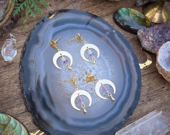 Puede incluir: Cuatro pares de pendientes dorados con forma de media luna con esmalte blanco y detalles de piedras preciosas moradas se exhiben sobre una rodaja de ágata gris y blanca. Los pendientes están colocados sobre una superficie de madera con cristales y otros objetos decorativos.