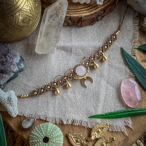 Puede incluir: Un collar de macramé hecho a mano con una piedra central de cuarzo rosa y detalles dorados. El collar presenta un dije de media luna y cuentas colgantes en forma de lágrima. Cristales y elementos decorativos adicionales están dispuestos alrededor del collar.