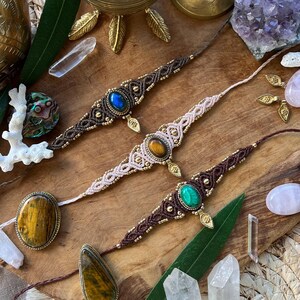 Peut inclure: Trois bracelets en macramé avec des pierres précieuses bleues, jaunes et vertes. Les bracelets sont exposés sur une surface en bois avec des cristaux, des feuilles et des objets décoratifs en laiton. Les bracelets sont faits à la main.