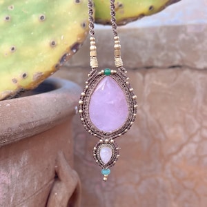 Peut inclure: Un collier macramé artisanal avec un grand pendentif en pierre précieuse rose clair en forme de goutte d'eau. Le pendentif est entouré d'un cordon marron tressé avec des perles dorées. Des pierres précieuses et des perles plus petites ornent le collier.