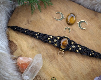 May include: A black macrame choker necklace with gold accents and a central oval tiger's eye stone. Additional tiger's eye stones and crescent moon charms are scattered around the necklace. The necklace is displayed on a wooden surface.
