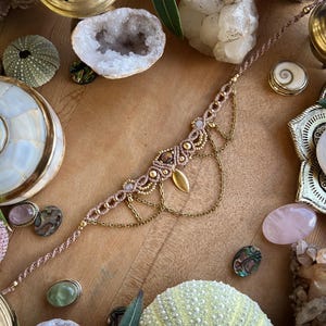 May include: A handmade macrame bracelet in shades of tan and gold, adorned with small beads and a gold leaf charm. The bracelet is displayed on a wooden surface, surrounded by crystals, shells, and decorative objects.