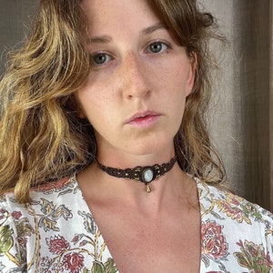 May include: A brown macrame choker necklace featuring a central oval, light blue stone and a small gold-coloured teardrop pendant. The necklace has gold-coloured beads and a woven design. The person is wearing a floral print top.