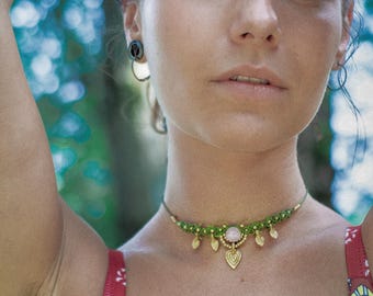 Puede incluir: Primer plano de una persona que lleva un collar choker de macramé verde con una piedra rosa y colgantes dorados en forma de corazón. La persona lleva una camiseta roja con un estampado floral. El fondo es verde borroso.