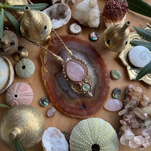 May include: A handcrafted necklace with a large rose quartz pendant, surrounded by intricate macrame work. The image also features various crystals, sea urchins, brass incense burners, and decorative shells, creating a bohemian aesthetic.