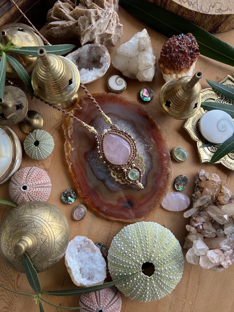 May include: A handcrafted necklace with a large rose quartz pendant, surrounded by intricate macrame work. The image also features various crystals, sea urchins, brass incense burners, and decorative shells, creating a bohemian aesthetic.