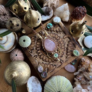 May include: A collection of bohemian-style jewelry and decorative items. A rose quartz pendant necklace is the focal point, surrounded by brass incense burners, crystals, sea urchins, and other natural elements. The color palette includes gold, pink, green, and brown.