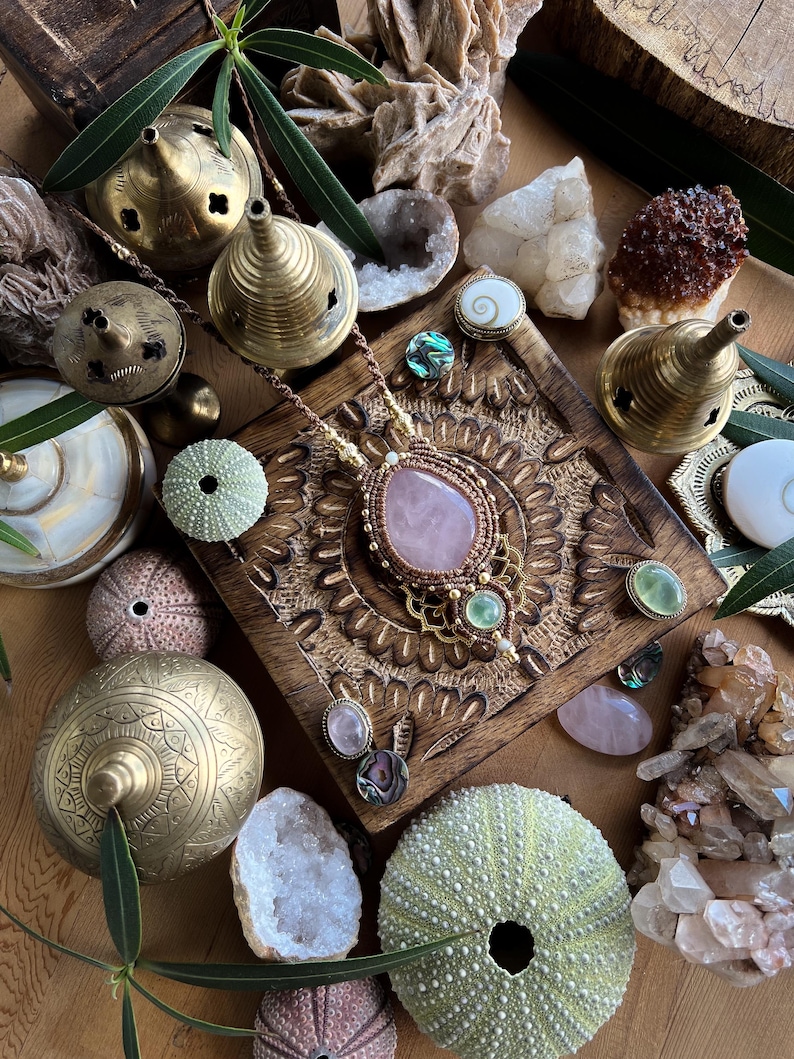 May include: A collection of bohemian-style jewelry and decorative items. A rose quartz pendant necklace is the focal point, surrounded by brass incense burners, crystals, sea urchins, and other natural elements. The color palette includes gold, pink, green, and brown.