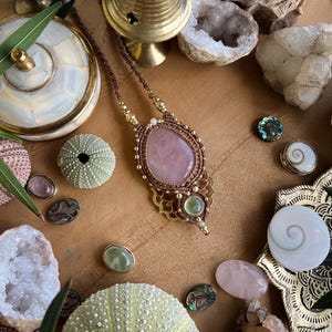 May include: A handcrafted macrame necklace with a large, oval rose quartz pendant. The necklace features a brown cord, gold accents, and green and clear gemstones. The image also includes decorative shells, crystals, and other natural elements.