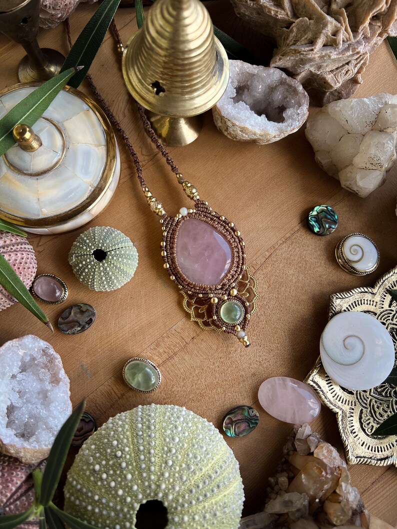 May include: A handcrafted macrame necklace with a large, oval rose quartz pendant. The necklace features a brown cord, gold accents, and green and clear gemstones. The image also includes decorative shells, crystals, and other natural elements.