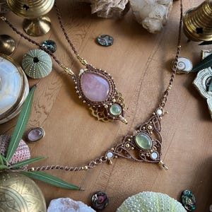 May include: Two handmade macrame necklaces with gemstone pendants. One features a large oval rose quartz, the other a green gemstone. Both are brown with gold accents and white beads. The necklaces are displayed with decorative objects.
