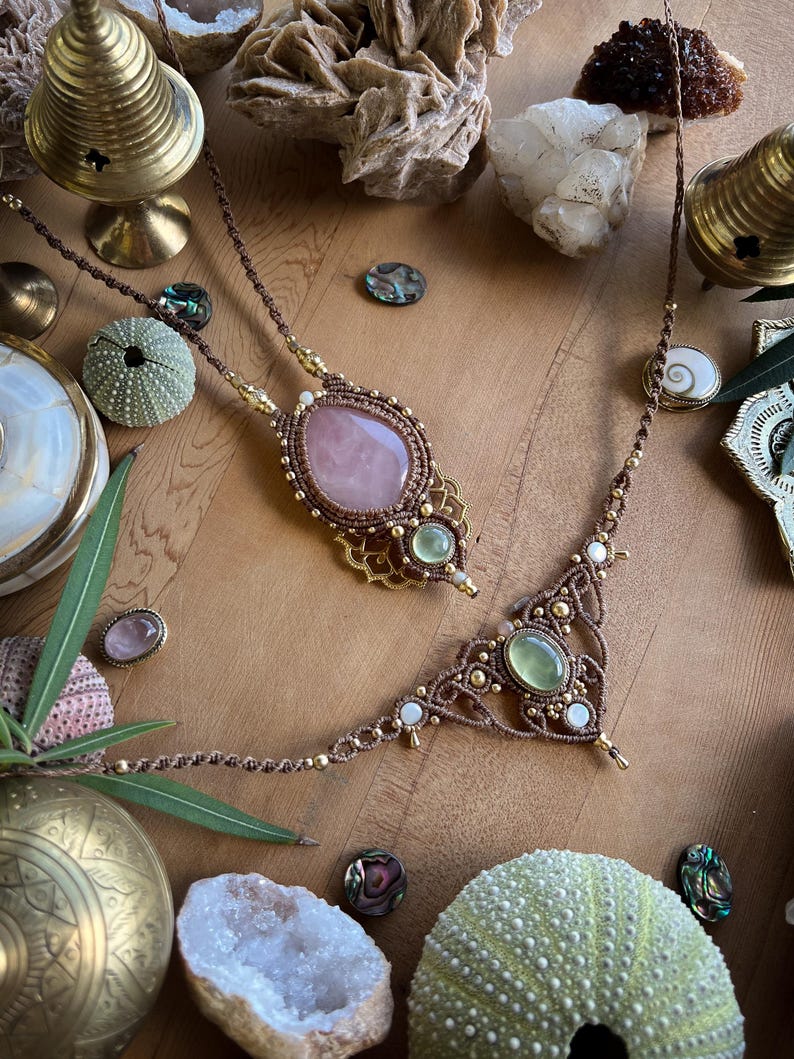 May include: Two handmade macrame necklaces with gemstone pendants. One features a large oval rose quartz, the other a green gemstone. Both are brown with gold accents and white beads. The necklaces are displayed with decorative objects.
