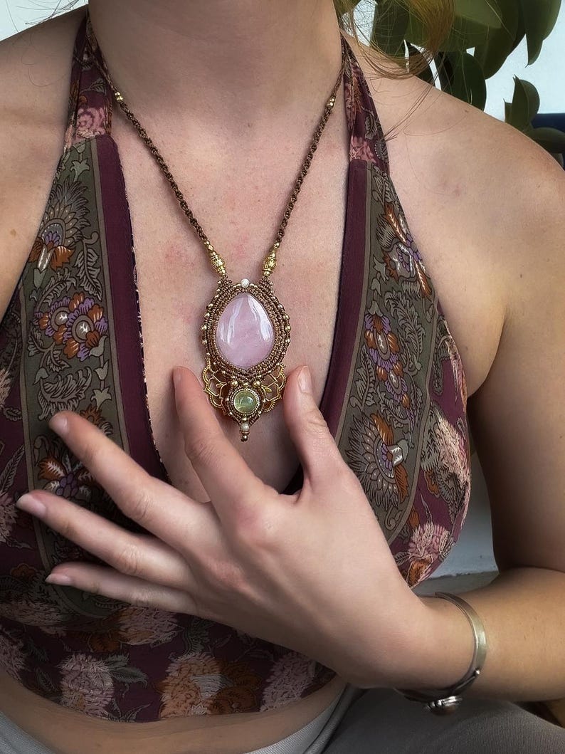 May include: A handcrafted necklace featuring a large, teardrop-shaped rose quartz pendant. The pendant is encased in a gold-colored, intricately woven setting, with a smaller green stone below. The necklace has a brown cord.