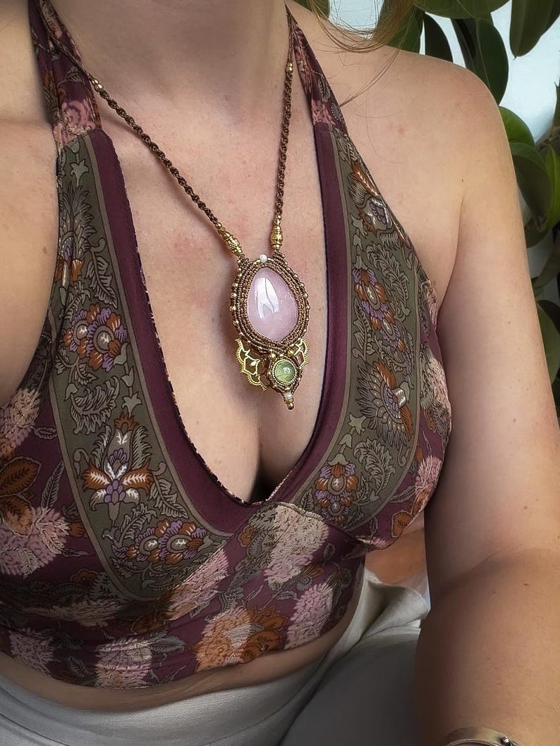 May include: A handcrafted necklace featuring a large, teardrop-shaped rose quartz pendant. The pendant is intricately wrapped with brown cord and accented with gold-colored metal details. The necklace is worn over a patterned halter top.