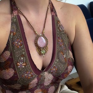 May include: A handcrafted necklace featuring a large, oval rose quartz gemstone pendant. The pendant is set in a macrame design with gold-colored accents and a smaller green stone. The necklace is worn with a halter top with a floral pattern.