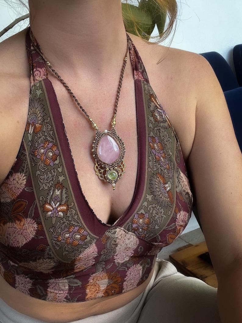 May include: A handcrafted necklace featuring a large, oval rose quartz gemstone pendant. The pendant is set in a macrame design with gold-colored accents and a smaller green stone. The necklace is worn with a halter top with a floral pattern.
