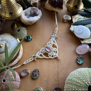 May include: A handmade macrame necklace with a pink gemstone center and gold accents. The necklace is surrounded by crystals, seashells, and small brass bells. The background is a wooden surface.