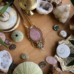 May include: A handcrafted necklace with a large, oval rose quartz pendant, surrounded by intricate macrame detailing. The necklace is displayed with various crystals, sea urchins, and decorative elements on a wooden surface, creating a bohemian aesthetic.