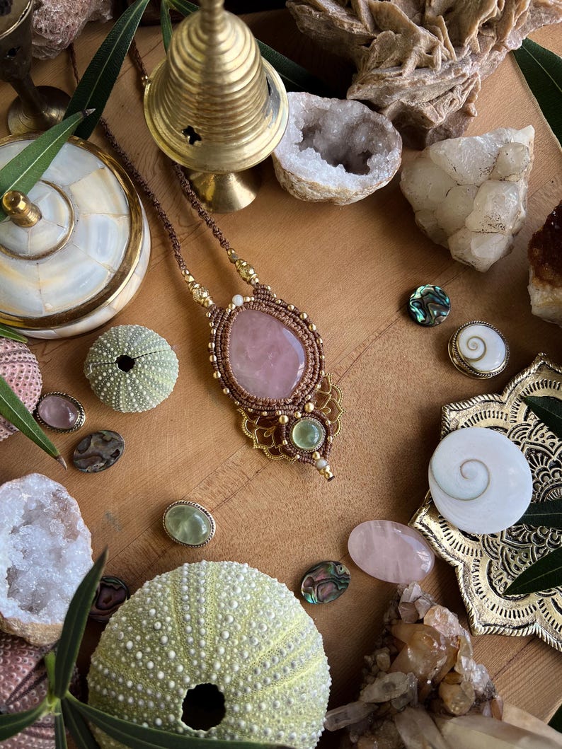 May include: A handcrafted necklace with a large, oval rose quartz pendant, surrounded by intricate macrame detailing. The necklace is displayed with various crystals, sea urchins, and decorative elements on a wooden surface, creating a bohemian aesthetic.