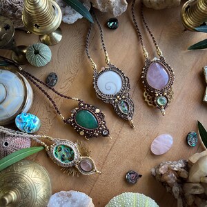 May include: A collection of handcrafted macrame necklaces with various gemstone pendants, including a white spiral shell, a green stone, and a pink rose quartz. The necklaces are displayed on a wooden surface with decorative elements like sea urchins and crystals.