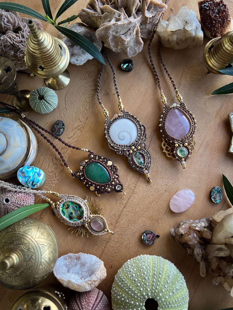 May include: A collection of handcrafted macrame necklaces with various gemstone pendants, including a white spiral shell, a green stone, and a pink rose quartz. The necklaces are displayed on a wooden surface with decorative elements like sea urchins and crystals.