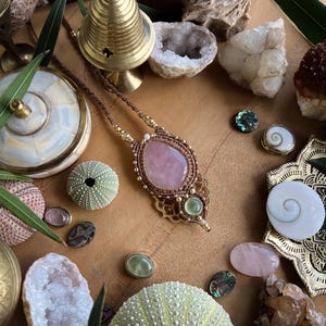 May include: A handcrafted necklace with a large, oval rose quartz pendant, accented with gold-colored metal and brown cord. The image also features various crystals, sea urchins, and decorative objects on a wooden surface, creating a bohemian aesthetic.