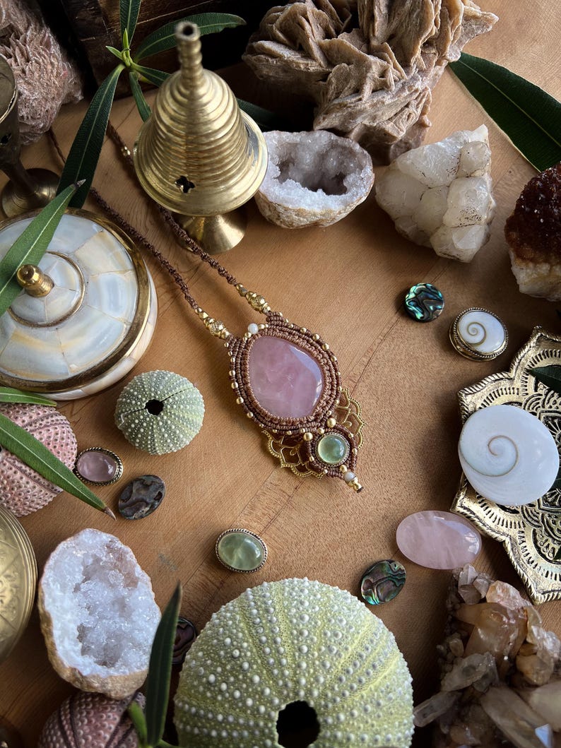 May include: A handcrafted necklace with a large, oval rose quartz pendant, accented with gold-colored metal and brown cord. The image also features various crystals, sea urchins, and decorative objects on a wooden surface, creating a bohemian aesthetic.