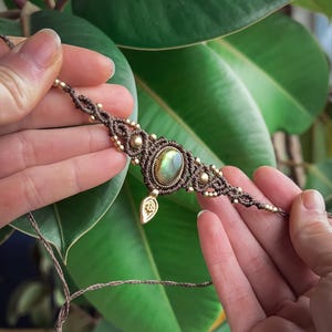 Op de afbeelding: Handgemaakte macramé armband met een centrale ovale edelsteen in groen- en goudtinten. De armband is gemaakt met bruin koord en versierd met kleine gouden kralen en decoratieve metalen elementen. Het heeft een bladbedel.