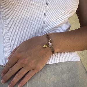 Op de afbeelding: Een bruine macramé armband met een roze edelsteen en goudkleurige details. De armband heeft een kleine gouden bedel. De armband wordt om een pols gedragen.