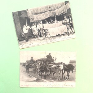 May include: Two vintage monochrome postcards. One shows people near a table with hanging items, possibly food. The other depicts a horse-drawn carriage on a road, pulled by several animals.