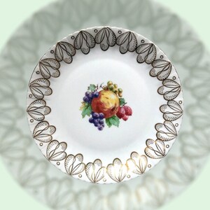 May include: A white ceramic plate with a gold rim and a decorative design of stylised hearts. The centre of the plate features a painted image of a fruit bowl with apples, grapes, and berries.