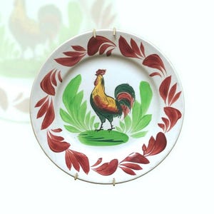 Pode incluir: Um prato de cerâmica branco com um design de galo vermelho e verde. O galo está em pé sobre uma base verde com folhas vermelhas ao redor da borda do prato.
