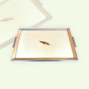 Può includere: Un vassoio d'argento con una superficie a specchio bianca e un piccolo disegno floreale al centro. Il vassoio ha un bordo decorativo e due maniglie.